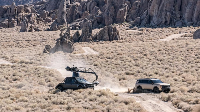 A color landscape photo of a scene being filmed for a movie. The setting is brown dry sagebrush with a windy dirt road. A gold colored SUV came racing down the road as evidenced by a dust trial. THe SUV is turning toward the right in the lower right side of the frame. Its rear tires are kicking up a lot of dirt. A modified black pickup truck, with a long camera boom, is close behind the SUV filming the action. In the background a many bizarre rock formations that look like clustered standing stones. 