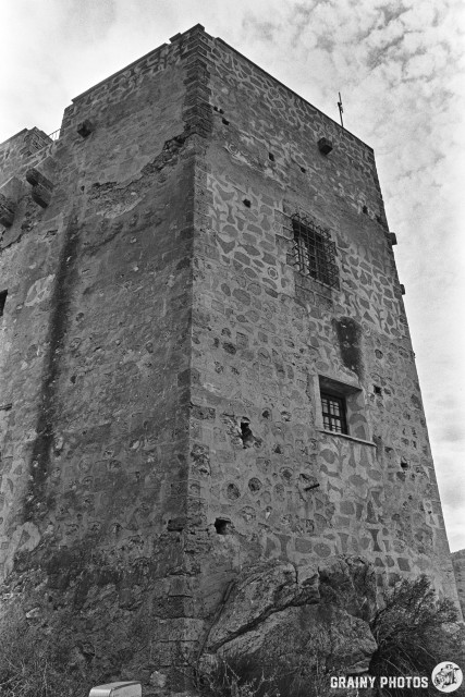 Black and white photo of the Castillo de Vélez de Benaudalla, an old stone tower with small windows and visible weathering. The sky above is partly cloudy, highlighting the building’s age.