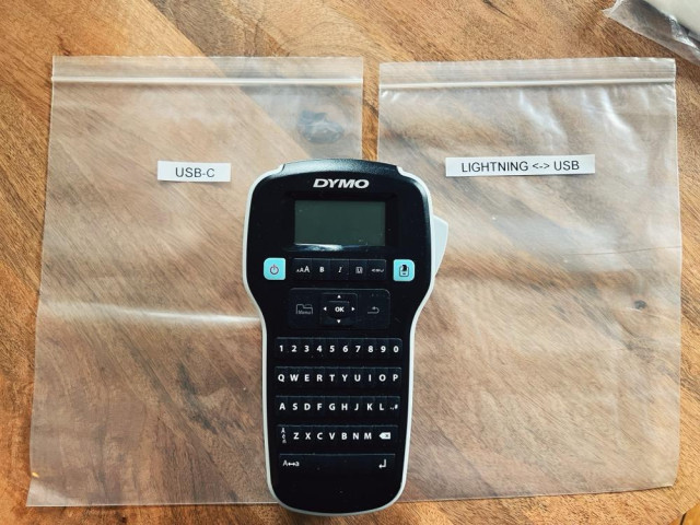 an overhead photo showing a Dymo labeler sitting on a table, in between two clear plastic bags. The one on the left has a white label that reads " USB-C" and the one on the right has a white label that reads "lightning <-> USB"