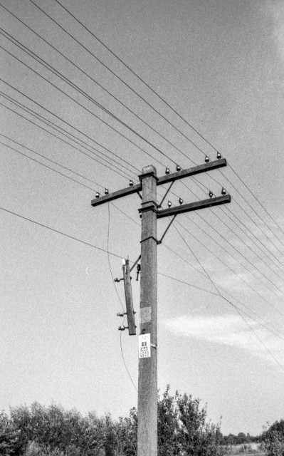 The black-and-white photograph shows a weathered concrete utility pole carrying a tangle of overhead telephone or power lines stretching diagonally across a bright, clear sky. The grainy texture of the image and the high contrast give it a distinctly analogue feel — likely shot on traditional film. A small metal sign attached to the pole marks a bicycle route, labeled **“R4 / R60.”** Behind the pole, low vegetation and trees line the horizon, suggesting a quiet rural or suburban landscape. The overall mood is minimalistic and slightly nostalgic, evoking the simplicity of everyday infrastructure in open countryside