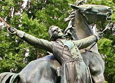  Bronze statue of a Confederate officer General Lloyd Tilghman on horseback, both arms outstretched with a sword in one hand, mouth open as if shouting, the horse rearing slightly behind him. The monument is set outdoors against a backdrop of green trees, evoking a dramatic, heroic pose typical of Lost Cause memorial sculpture.