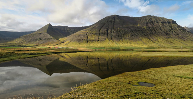 A colour photo of typical Icelandic mountains - craggy, glacial and cut by water. The strong sunlight is touching the green flanks of the mountains, and they're reflected in the still waters of the fjord.