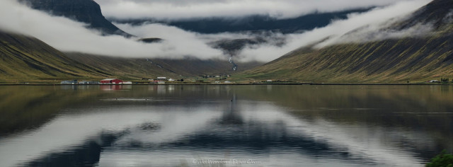 A colour photo of a body of water with mountains and a U-shaped valley in the distance. Buildings are scattered along the shoreline. The mountain flanks are coated with cloud but the peaks are still visible. The scene is reflected in the slightly ruffled water.