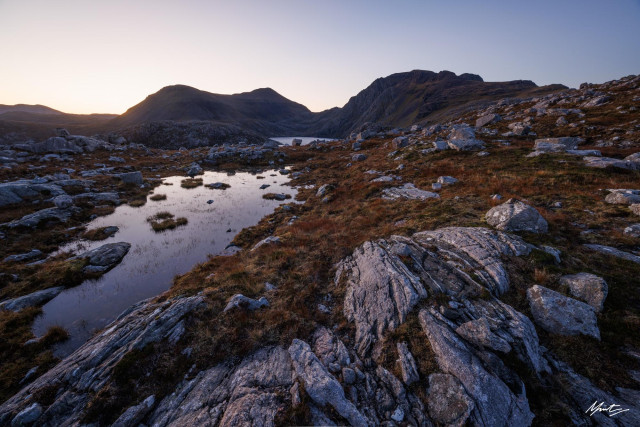 a photograph of the remote northwest scottish highlands, featuring a sweeping foreground of gneiss, autumnal coloured deer grass, a wee lochan on the right hand side. These elements all converge towards two mountains in the distance. The scene is illuminated by soft glowing pre-dawn light with clear skies above. 