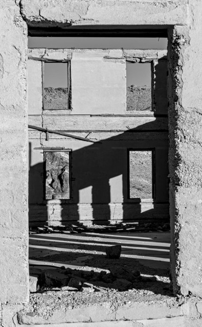 A black and white portrait photo looking through the large empty window frame of a long abandoned commercial building. All that stands of the building are the outer concrete walls. Looking through the window frame in the foreground you can see into the building and the opposite wall which reveals the building to be two stories with two empty window frames on the upper floor and two empty window frames on the bottom floor. A nearby rocky hillside is seen in the distance through the empty window frames. 