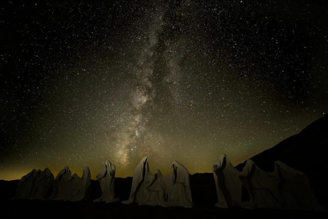 A color landscape photo of the night sky. The background of the image is the Milky Way Galaxy. The Galaxy is seen as a band of dense stars angling up vertically from the lower part of the frame to the top. There is skyglow along the horizon in the background. Skyglow is light reflected on dust particles from the sun that is far below the horizon. In the foreground is a sculpture of a series of ghostly caped figures on a platform. They are made to replicate the figures in Leonardo da Vinci's Last Supper painting. The figures are dimly lit under the starlight.