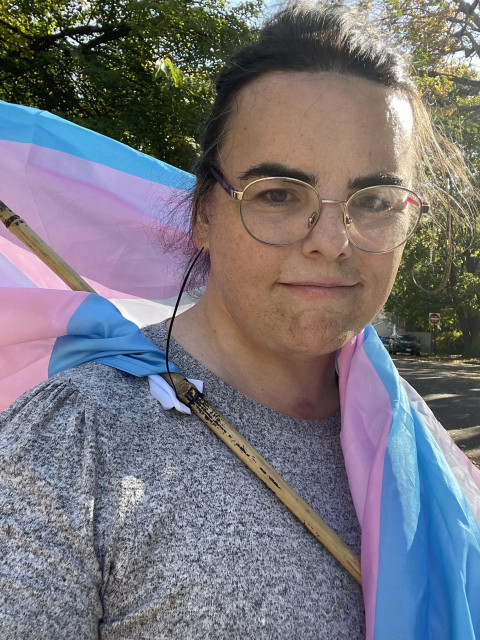 Me, in one of the worst selfies I've ever taken, dirty face and all.  A woman with black hair tied back into a bun, wearing glasses and a gray sweater.  The bamboo pole (one that holds a trans flag) crosses her chest; the trans flag drapes over one shoulder.  Trees and foliage are in the background.  