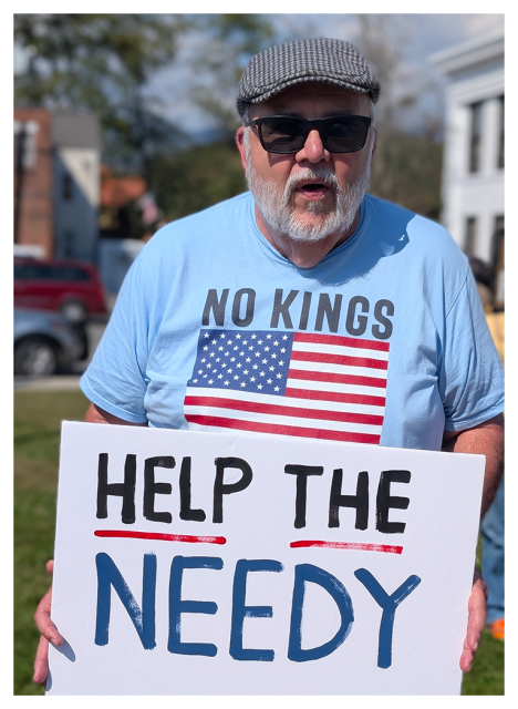 An older man with a white beard and sunglasses wears a gray flat cap and a blue NO KINGS shirt with an American flag. He holds a sign reading HELP THE NEEDY at a daytime outdoor event, with buildings and trees visible in the background.