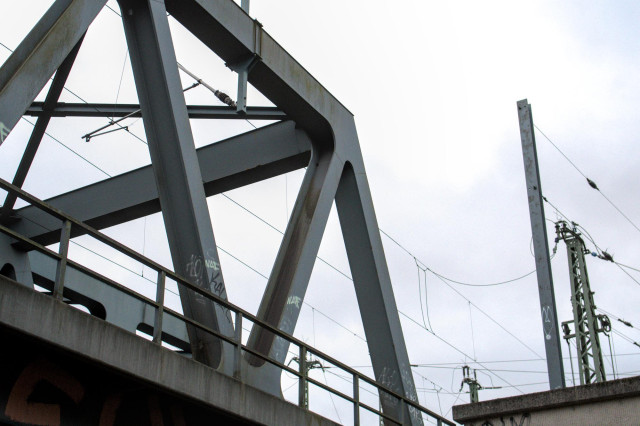 Stählerne Fachwerkbrücke mit Graffiti, darüber Stromleitungen und ein Strommast vor bewölktem Himmel.