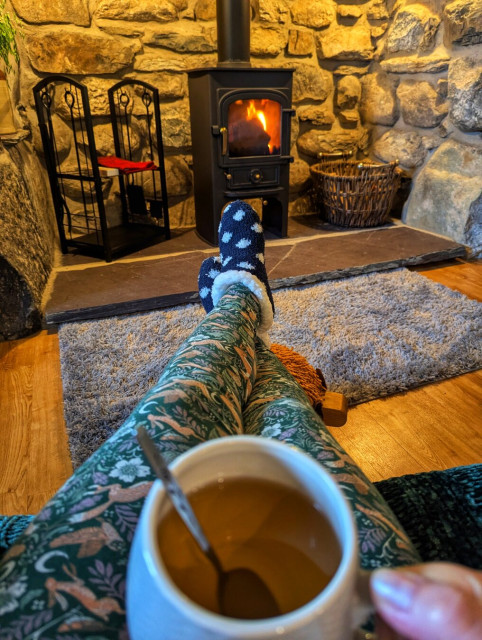 A cosy indoor scene featuring a person relaxing by a lit wood-burning stove. The individual is seated with their legs stretched out, wearing patterned green leggings and blue socks with white polka dots. They are holding a large mug of hot tea or coffee, with a spoon inside. The setting includes a rustic stone wall and wooden floor, with a grey rug placed in front of the stove. Beside the stove, there is a wicker basket and a set of fireplace tools. The atmosphere is warm and inviting, evoking a sense of comfort and relaxation.