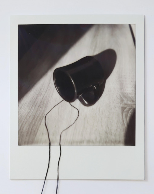 This Polaroid photograph depicts a black ceramic mug lying on its side on a wooden surface, illuminated by soft, directional light that creates gentle shadows. Two black threads are physically attached to the bottom edge of the instant photo, extending beyond its white border. These threads act as a visual continuation of the image — as if something from within the frame is spilling or flowing out into the real world. The composition blurs the line between the photographed and the tangible, evoking a quiet surrealism and a sense of transition between illusion and reality.
