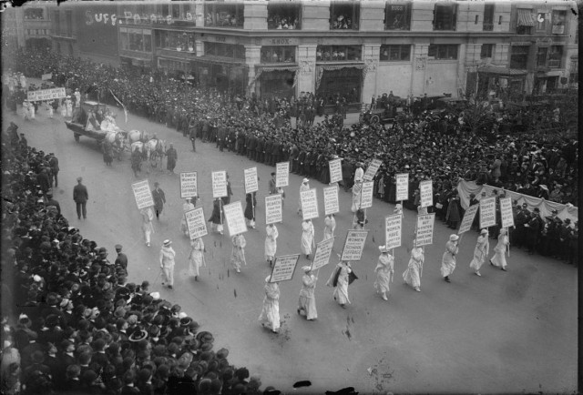 large crowds watch the parade. The front has 24 women all in white and carrying banners. Everyone is white.
