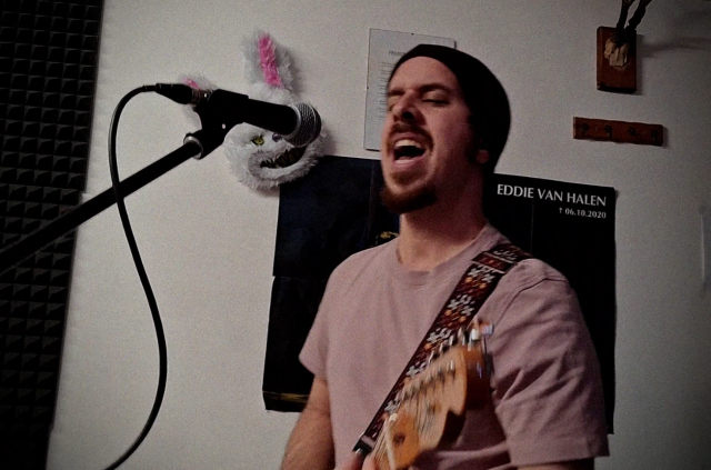A bearded guy in a beanie and T-shirt with a guitar, yelling into a microphone. He's in a room with white walls, a hunting trophy and a scary bunny mask on them.