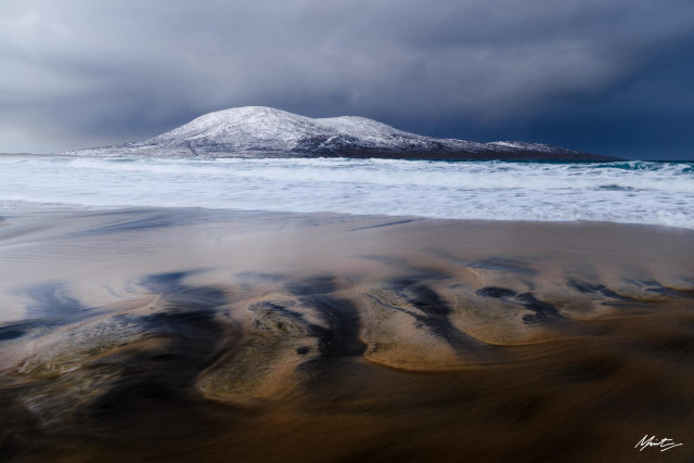 a snow capped hill sits centrally in the background of this coastal photograph, with streaks of dark peat in the foreground running like veins through the white sand. The ocean is tumultuous and frothy with winter rage. 