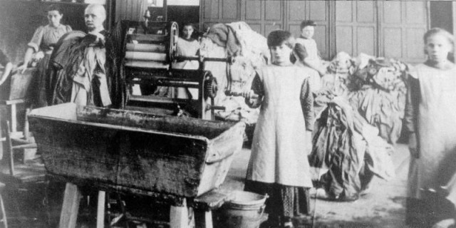A young girl standing by a huge mangle, with mounds of laundry behind her. Other workers are in the background.

