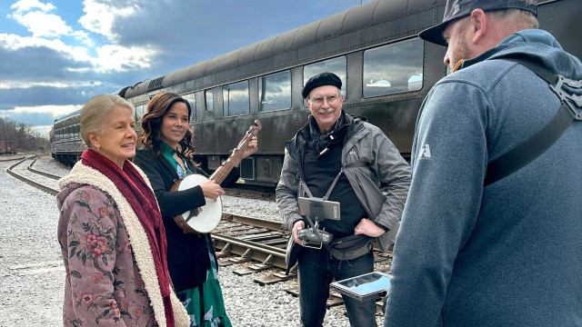 Rhiannon Giddens (second from left, with a banjo) stands outdoors near a train with crew members on the set of My Music with Rhiannon Giddens, Season Two.