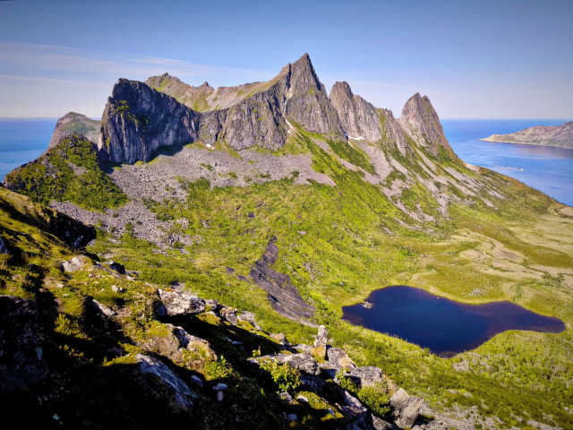 The image depicts a stunning mountainous landscape with a prominent peak in the center, characterized by sharp, jagged rock formations and steep slopes. The mountains are covered with patches of green vegetation, indicating a mix of rocky and vegetated terrain. A serene, dark blue lake is nestled in the foreground, surrounded by lush greenery, adding a contrasting element to the rugged mountains. The background features a clear blue sky and a body of water, possibly an ocean or a fjord, which extends to the horizon. The sunlight casts shadows on the mountains, highlighting their texture and depth. The overall scene is a picturesque representation of a natural, untouched environment, showcasing the beauty of the landscape.
