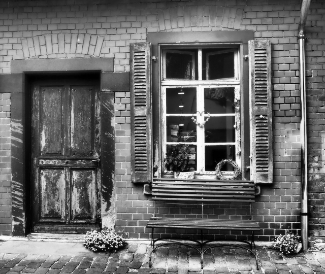 eine Aufnahme von einem kleinen Backsteingebäude. Man sieht eine alte Holztür die als Eingang fungiert. Rechts daneben ist ein hohes großes weißes Sprossenfenster. Unter dem Fenster steht draussen eine Bank zum hinsetzen.. 