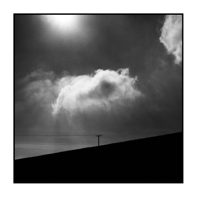 Square black-and-white photograph. A large, bright cloud dominates the upper center of the frame, illuminated by diffuse sunlight from the top left. Below, a dark diagonal slope cuts across the image from lower left to upper right. On the ridge line, a single utility pole with two faint wires stands against the sky, small and silhouetted.