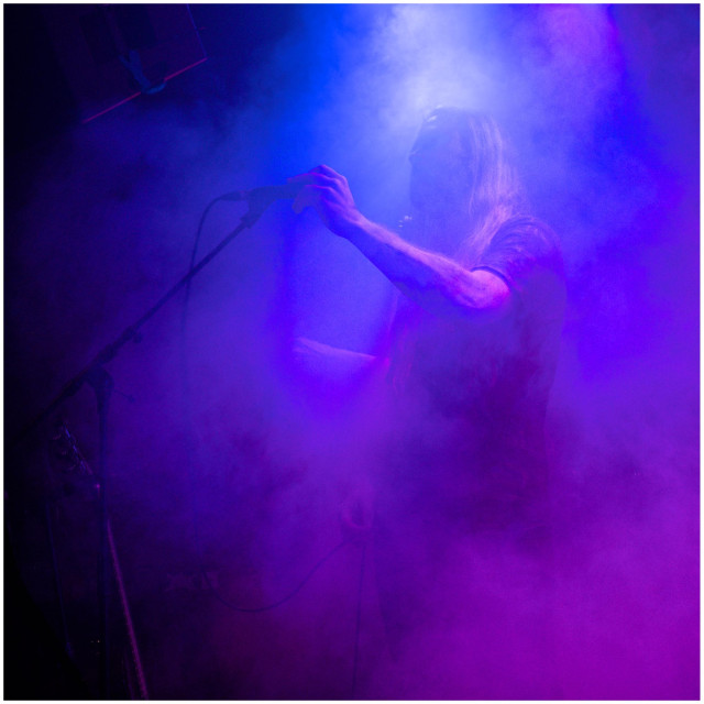 Person mit langen Haaren steht im Bühnennebel und greift nach einem Mikrofon vor blau-violettem Licht