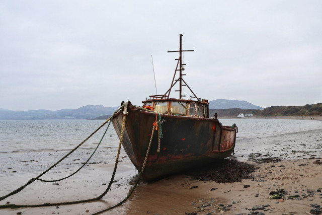 A weathered, rust-coloured fishing boat resting on a sandy shoreline at low tide. The boat, showing signs of age and exposure to the elements, is anchored to the shore with thick ropes. Its tall mast and rigging are visible against a muted, overcast sky. In the background, rolling hills and a distant coastline create a serene, desolate seascape. The scene evokes a sense of quiet abandonment and the passage of time.