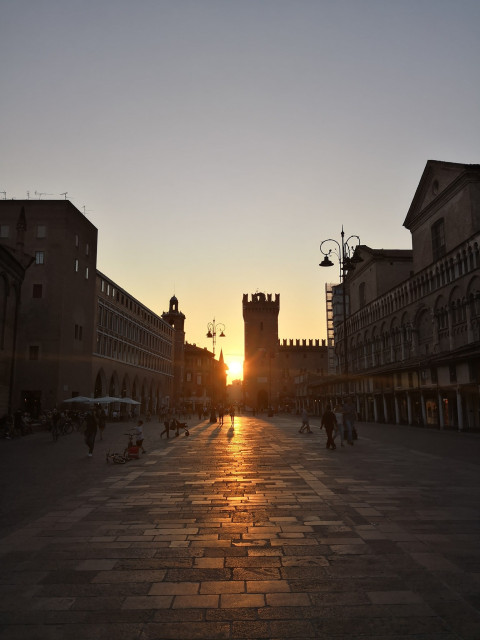 Sunset in an Ferrara's Piazza Trento e Trieste, where the 'kiss of the sun' bathes the cobbled path in a warm, golden light. Silhouettes of historic buildings and a classic watchtower stand against the glowing sky. Pedestrians become dark figures in the sun's embrace, casting long shadows that blend with the outlines of ancient architecture. The tranquil moment captures the romance of the day's end, as the sun's farewell touch graces the heart of the city with a fleeting, amber radiance.