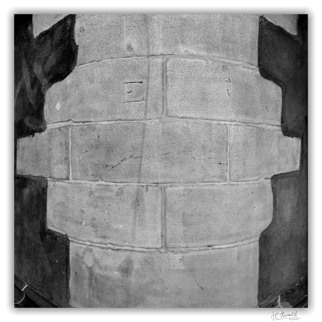 Square black-and-white image of a round corner of Frankfurt Cathedral. The corner is made of large light grey stones and merges into the walls with dark plaster. At the top, to the left of the centre, a large E is engraved into one of the grey stones. 