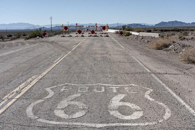 A color landscape photo of a highway going from the bottom of the frame to the top. The road is grey with a solid yellow line on the left and to the left of the solid line is a dashed line. Ahead there are barriers that say, "Road Closed." It's a desert landscape with a range of mountains low on the horizon. The sky is clear and blue. 