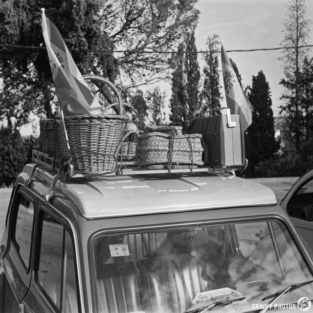 A vintage car with wicker baskets and decorative flags on its roof, set against a backdrop of trees in black and white. The scene conveys a nostalgic, leisurely atmosphere, possibly for a picnic or outdoor excursion.