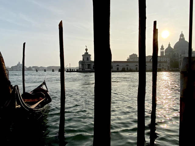 Colour photo. In the centre of the image, a wooden mooring post, like a deep black line running from top to bottom of the frame, at the head of a V-shape of four other posts going back into the silvery gently rippled water of a wide canal. On the left of the frame, a moored black gondola. In the mid ground is the Salute basilica, with its two majestic domes, with the setting yellow-haloed sun just above it, making the building shimmer grey in the hazy light, and leaving a streak of golden flecks on the water. Further back, the low skyline of Giudecca island in the distance, and pale grey sky above. 