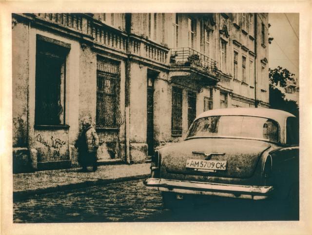 This lith print captures a quiet street scene in the old part of Lviv. The composition features a vintage car parked along a cobblestone road, its dusty surface echoing the texture of the worn building façades. A solitary figure stands near the wall, wrapped in a heavy coat, adding a sense of human melancholy and timelessness to the frame. The print’s warm, gritty tones and soft contrast typical of the lith process evoke a nostalgic, almost cinematic atmosphere — as if the city itself is suspended somewhere between memory and dream