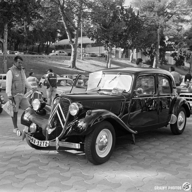 A classic black Citroën car parked at a classic car show, with people mingling in the background. Trees provide shade as a man in a jacket stands beside the vehicle, contributing to a nostalgic automotive atmosphere.