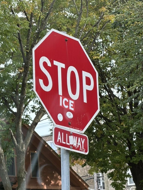 A red octagonal stop sign is shown with the word “STOP” in large white letters and the word “ICE” in smaller white letters below it. Beneath the stop sign, there is a smaller sign that reads “ALL WAY,” although some of the letters are partially covered or damaged. The sign is mounted on a metal pole and surrounded by green trees and residential houses in the background.