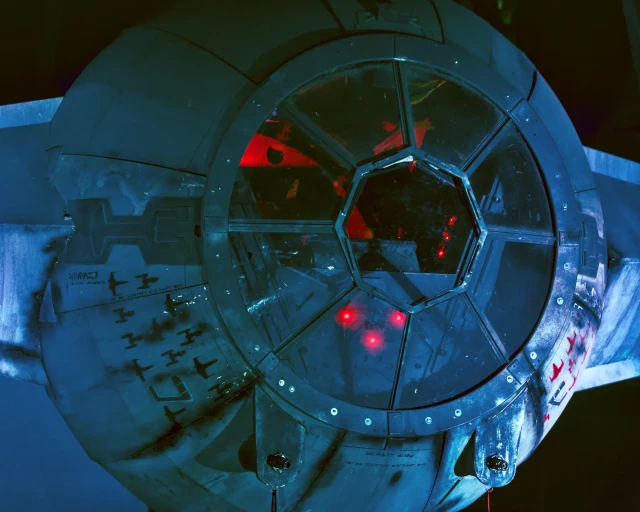 Nahaufnahme der Pilotenkanzel eines Tie-Interceptors. Aus dem Inneren leuchten rote Lampen, der hinter Teil der Kanzel ist rot erleuchtet. Die runde Pilotenkanzel ist übersät mit Macken, Brandspuren und Kratzern. 
Links und rechts an den Seiten sind ein Dutzend Sticker abgeschossener Rebellen-Kampfraumschiffe zu erkennen. 
Kleines Detail: An beiden Laserkanonen hängen noch die Sicherungsstifte. Folglich befindet sich der Tie-Interceptor in einem Hangar. 
. 
Close-up of the cockpit of a Tie Interceptor. Red lights shine from the inside, and the rear of the cockpit is also illuminated in red. The round cockpit is covered with dents, burn marks and scratches. 
A dozen stickers from downed rebel combat spacecraft can be seen on the left and right sides. 
Small detail: the safety pins are still attached to both laser cannons. This indicates that the Tie Interceptor is located in a hangar. 
. 
