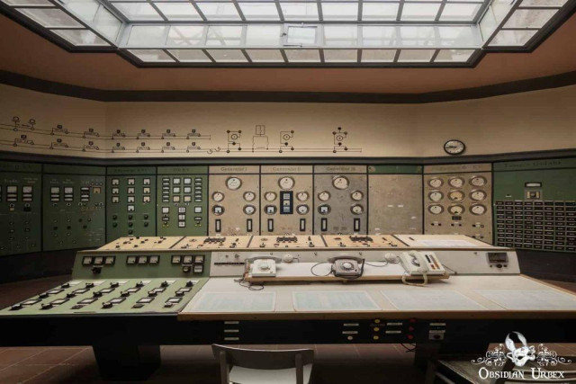 A vintage industrial control room with large control panels, numerous gauges, dials, and switches, and a central desk with phones under a skylight ceiling.