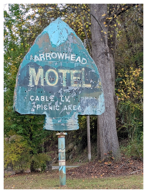 	
A weathered, arrowhead-shaped metal sign reads “Arrowhead Motel, Cable T.V., Picnic Area.” The paint is faded and peeling. The sign stands in front of tall trees with green and yellow leaves, and theres grass and brush in the background.