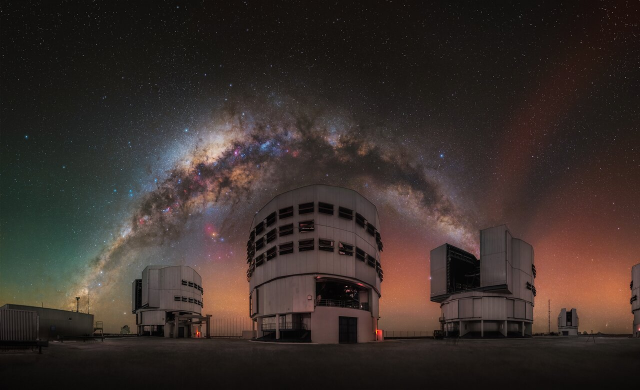 An observing site, with three large domes in the centre and the arching Milky Way above them. To the left is a long building, and to the right is a smaller dome. A fourth, larger dome is hidden on the right side. The horizon exhibits a faint coloured glow: green on the left and red on the right