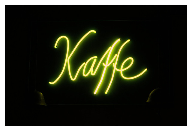 En liggande bild på en gul neonskylt med texten ”Kaffe” mot en svart bakgrund. I de nedre hörnen av skylten faller lite ljus på två armé-hjälmar.