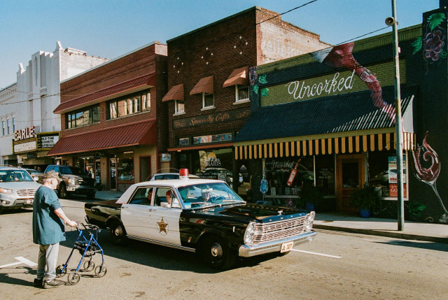 A photo of early 1960s Ford police car driving on the street of a small town.  There is an older guy with a rolling walker/buggy waiting to cross the street.