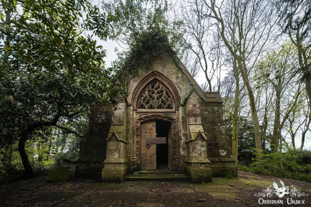 An abandoned, gothic-style stone crypt stands in a forest clearing. The crypt has a tall, narrow roof, a large pointed arch doorway with a partially open wooden door, and a decorative circular window above the entrance. The building is overgrown with ivy and vines, and is surrounded by trees with bare branches. The ground is covered in dirt and fallen leaves.