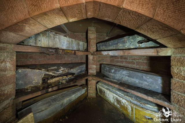 The image shows the interior of a brick and stone crypt or mausoleum. Several metal coffins or caskets are visible, resting on wooden and stone shelves. The space appears to be old and in a state of disrepair, with water on the floor and signs of decay