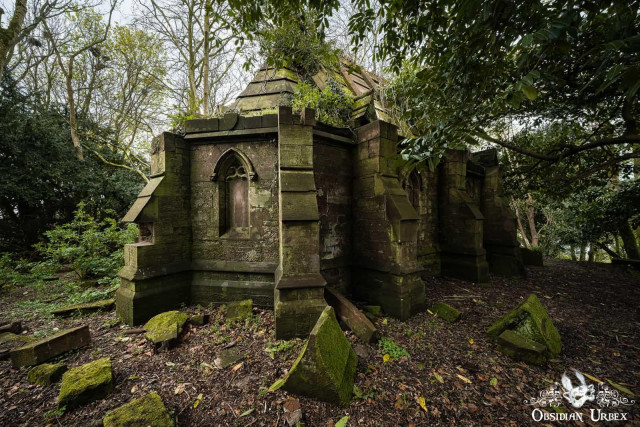 The stone building is in a state of disrepair, with moss and vegetation growing on its walls and roof. The mausoleum is surrounded by trees and fallen stones, giving it a gothic and overgrown appearance
