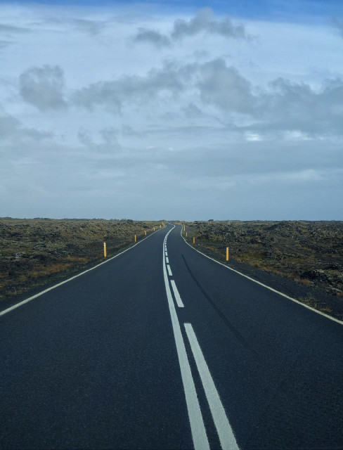 A photo showing a road with new tarnac and white center lines stretching into the distance through a rocky, barren landscape under a cloudy sky.

