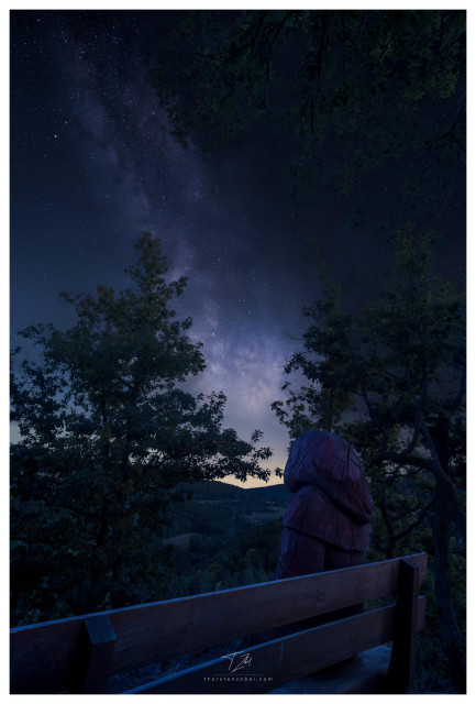 A wooden man wearing a hood sits on a bench at night, looking out over a valley. Above the valley, the Milky Way is clearly visible, framed by oak trees in the foreground.