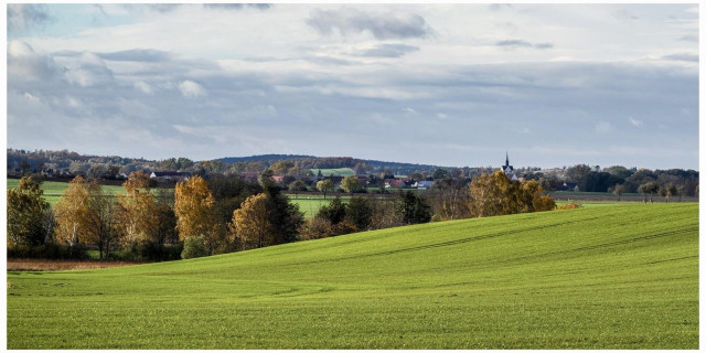 A scenic landscape featuring rolling green hills, scattered trees with autumn foliage, and a distant village with a church steeple against a cloudy sky.