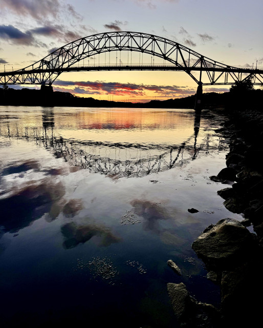 Photo of bridge over calm reflective water. Sunset in background. Dark clouds overhead and in reflections.

Sagamore Bridge, Massachusetts.