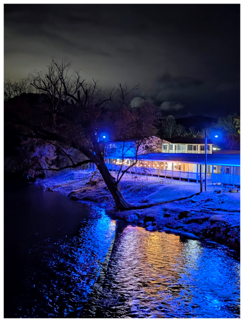 ai-assisted alt text:

	
A warmly lit building sits by a river at night, surrounded by trees under a cloudy sky. Blue lights illuminate the water’s edge and trees, reflecting brightly on the rippling river. The scene feels quiet and serene with dramatic lighting contrasts.