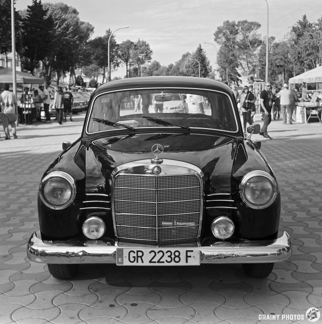 A vintage black Mercedes-Benz sedan is parked prominently, showcasing its distinctive grille and classic design, with a bustling outdoor event and spectators in the background. The scene captures a nostalgic automotive gathering.