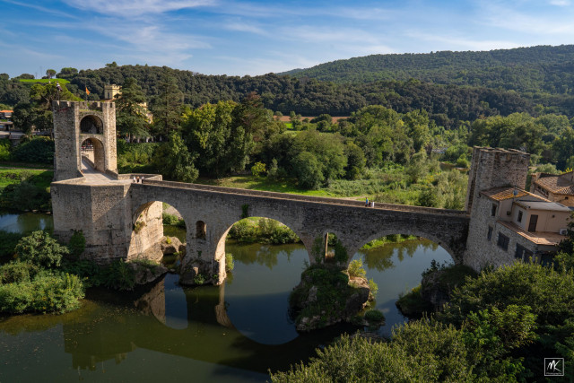 Color photo of an old stone bridge with three arches and a tower spanning a river, with green hills in the background 