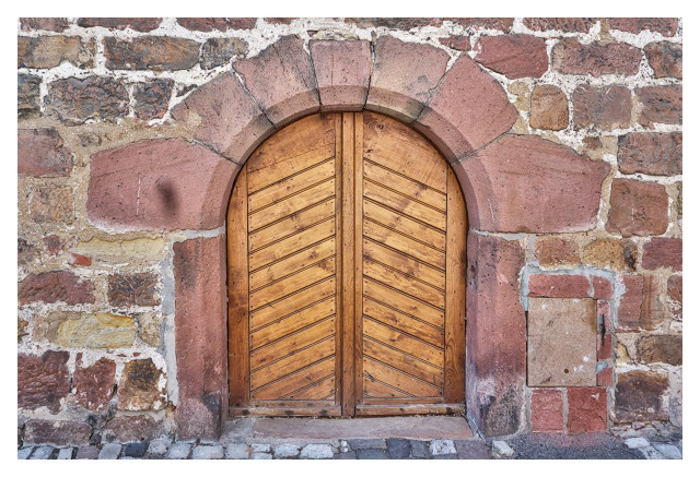 A close-up photograph of an old wooden double door set into a rustic stone wall. The door is arched at the top, framed by a rounded arch made of reddish-pink sandstone blocks. The door panels are constructed from vertical wooden planks arranged in a chevron or herringbone pattern, pointing inward toward the center seam where the two halves meet. The wood appears weathered, with a warm brown tone and visible grain. Surrounding the door, the wall is composed of irregularly shaped stones in shades of red, brown, beige, and gray, mortared together with some white plaster visible in the joints. To the right of the door, there's a small rectangular stone slab or cover embedded in the wall, possibly a access panel or old mailbox slot. The ground in front is paved with uneven cobblestones. The overall scene evokes a historical or medieval European building, with signs of age like dirt, cracks, and slight discoloration on the stones and wood.